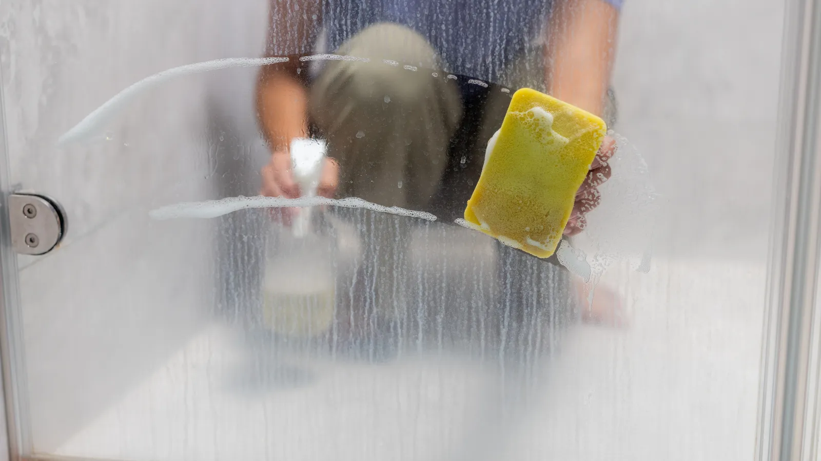 Close view of someone on other side of glass shower door, using yellow sponge and soap to scrub off hard water stains.