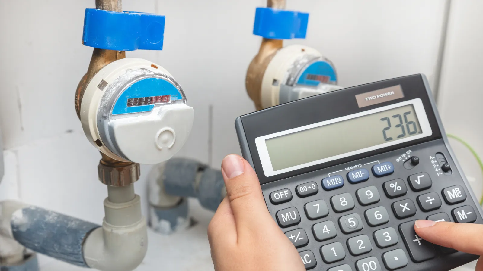 Close-up of a person's hands using a calculator while standing in front of a water meter installed in a white cement basement wall.