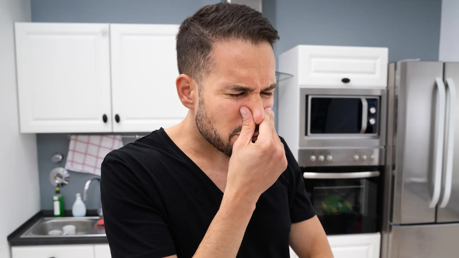 Young man standing in a kitchen, pinching his nose to block out a bad odor.