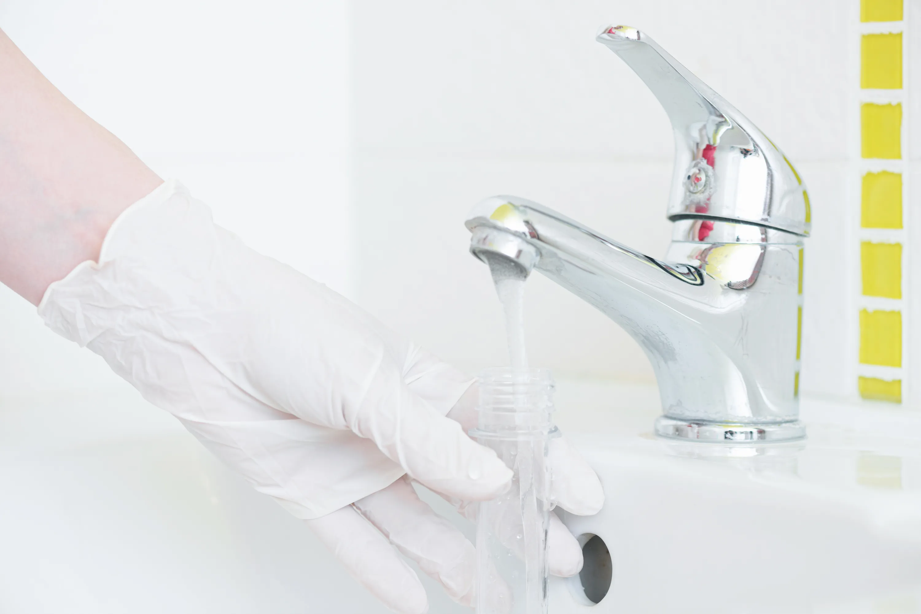 A person's hand in a white rubber glove, holding a white plastic tube under a silver bathroom faucet running water.